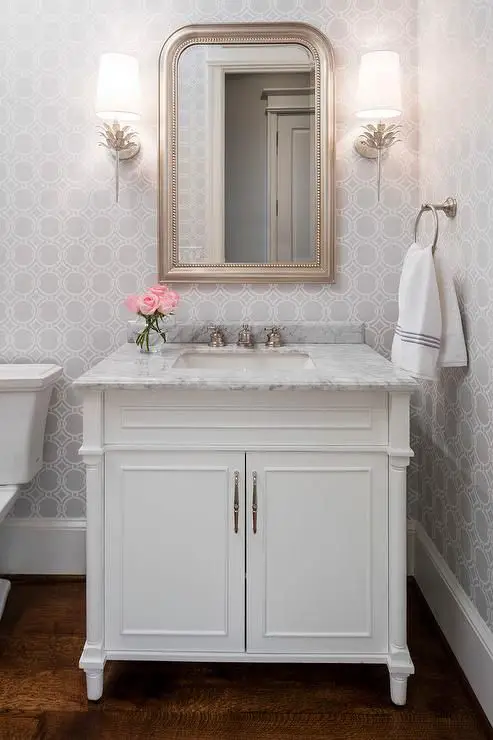 White and gray powder room features walls clad in gray geometric wallpaper lined with a white single washstand topped with carrera marble placed under a silver leaf beaded mirror illuminated by Worlds Away Delilah Silver Wall Sconces.: 