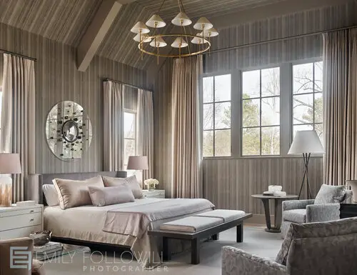Beige designed bedroom with double bed, chair at the bottom of the bed, chandelier, mirror on the wall above the bed and windows on the side of the bed