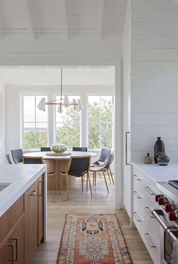 View from kitchen to dining room with table and chairs around it with big windows behind