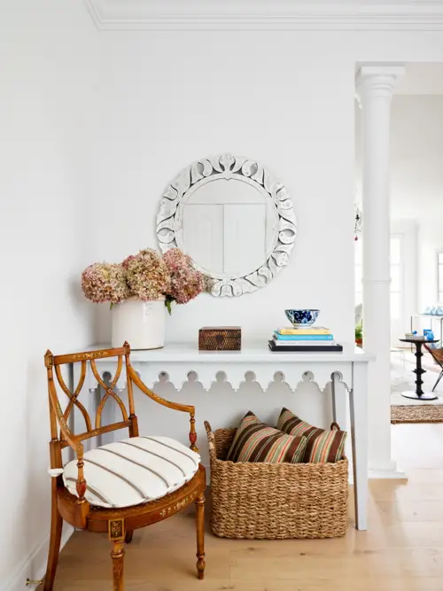 Part of the bright designed living room with chair, table and pillows in the basket below the table, and a mirror on the wall above the table with flowers and books