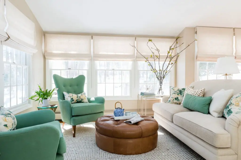 Living room with wide windows and beige sofa with colorful pillows on it, and a couple of green armchairs, with coffee table in the middle