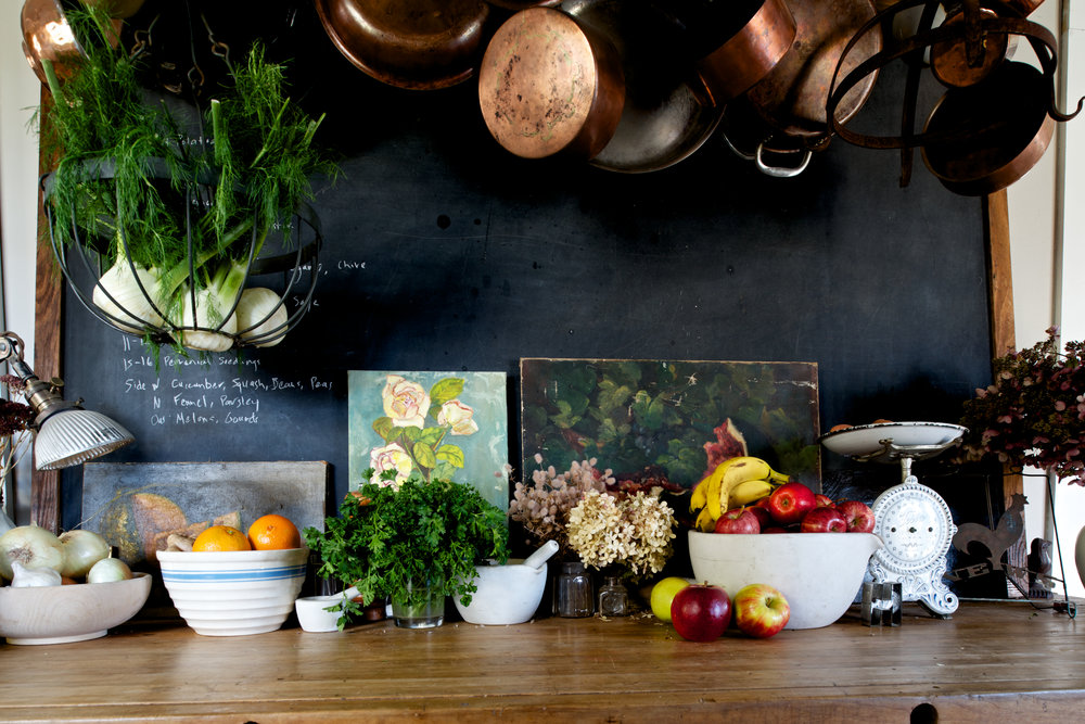 Table with bowls of fruits, parsley, flowers and vegetable with pictures behind the bowls on the dark wall