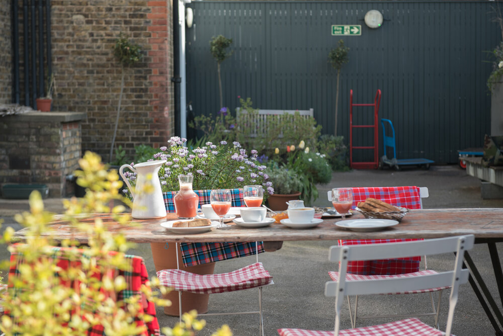 Table with breakfast served on it with chairs around in the backyard with plants in pots