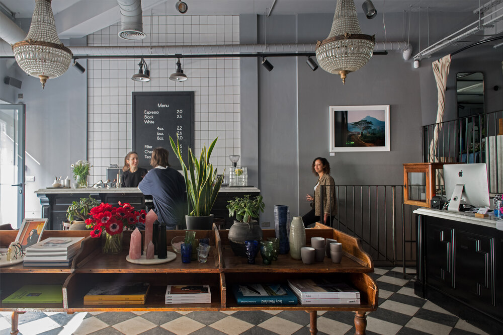 Store with table that has candles, vases and books on it with a counter in behind and woman serving coffee to the costumer while the other woman's entering the room